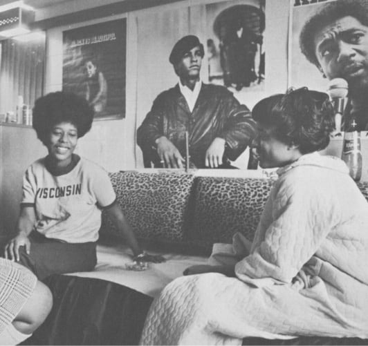 Two Black students sitting on a couch in a residence hall with posters of Black activists displayed behind them.