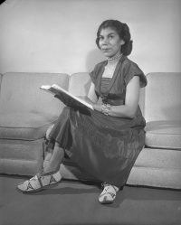 Geraldine Harvey sitting cross-legged with a book wearing a dress and moccasins.