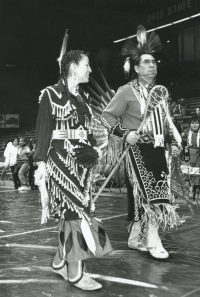 Two tribal members walk side by side in the UW–Madison Field House dressed in full Native regalia.