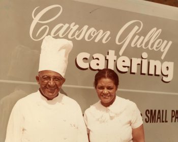 Carson Gulley and his wife, Beatrice, smiling and posing in front of their catering vehicle that reads "Carson Gulley catering". Both are dressed in their chef uniforms.