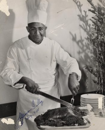 Portrait of Carson Gulley slicing into one of his culinary creations, a giant roasted bird. The chef is smiling, a christmas tree is behind him, his tall chef's hat extends out of the top of the picture. The image is autographed.
