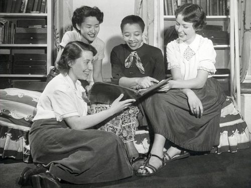 Four women sit together smiling and looking at a book together. They are a diverse group.