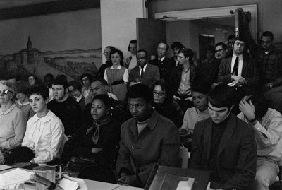 A crowded room of people, Black and white, inside a room. The foreground has two rows of seated solemn people, and more people are standing at the back of the room in the doorway.