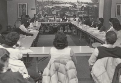 A large conference room on campus shows full attendance of people seated with jackets on their chairbacks. A large mural fills the wall in the background.