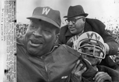 Photograph of Lewis “Les” Ritcherson, Milt Bruhn, and a football player cheering and holding one another at a football game with crowds in the background.