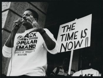 Charles Holley speaking into a mic about racism on campus wearing a “Black by Popular Demand - University of Wisconsin Madison” crew neck next to a sign which reads “The Time is Now!!”