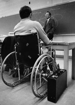 A professor lectures in front of a chalkboard while a student in a wheelchair listens, a briefcase by his side.