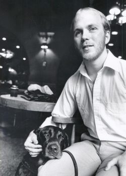A student in white shirt and light pants seated with his dog's head resting on his knee. Dark lighting behind them.