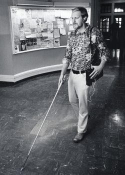 A man in white pants and a patterned shirt walks with a cane through a hallway on campus. He has a tape recorder attached to his waist and his hand is on it. A busy bulletin board is behind him.