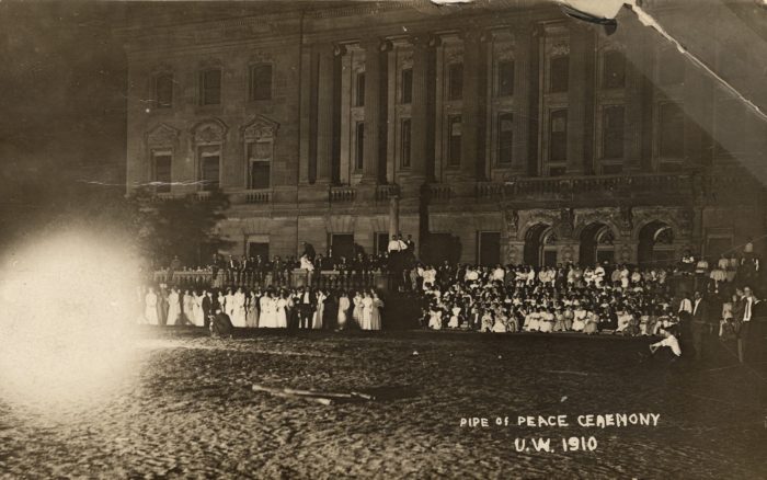 A large crowd stands in front of the Wisconsin Historical Society building at the Pipe of Peace ceremony. It appears to be nighttime and there is a large photo flare in the lower lefthand corner obliterating part of the image.