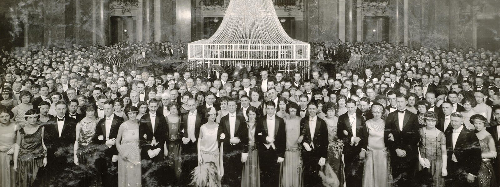 Several hundred students gathered around the rotunda at the Wisconsin State Capitol during the 1924 junior prom. They are dressed in formal attire beneath a crystal chandelier.