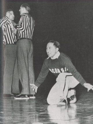 Toru Iura is on one knee during a cheer, smiling, with hands outspread. Two referees in discussion behind him.