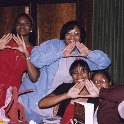Members of Delta Sigma Theta Sorority, Inc. posing with one of their signature hand signs sitting around a table.