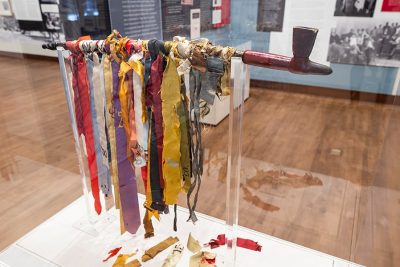 A wooden pipe sits on a stand and displays numerous ceremonial ribbons of different lengths and colors that drape over it.