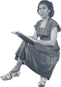 Geraldine Harvey sitting cross-legged with a book wearing a dress and moccasins.